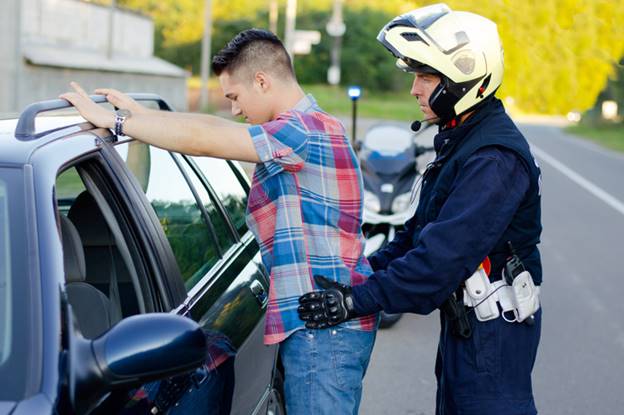 A man in a plaid shirt is being searched by a police officer. The search is taking place beside a car on the side of a road.
